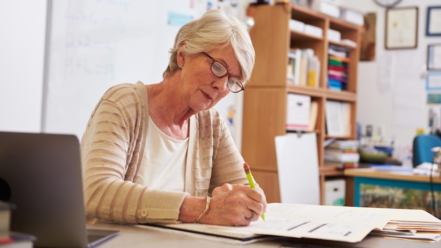 An older woman sits at her desk at the front of a class and writes on an exam booklet.