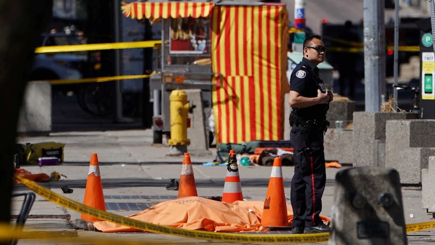 A police officer behind police tape stands next to a victim, who is covered.