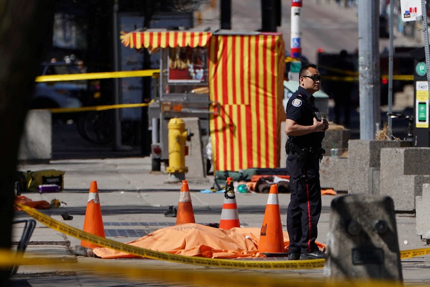 A police officer behind police tape stands next to a victim, who is covered.