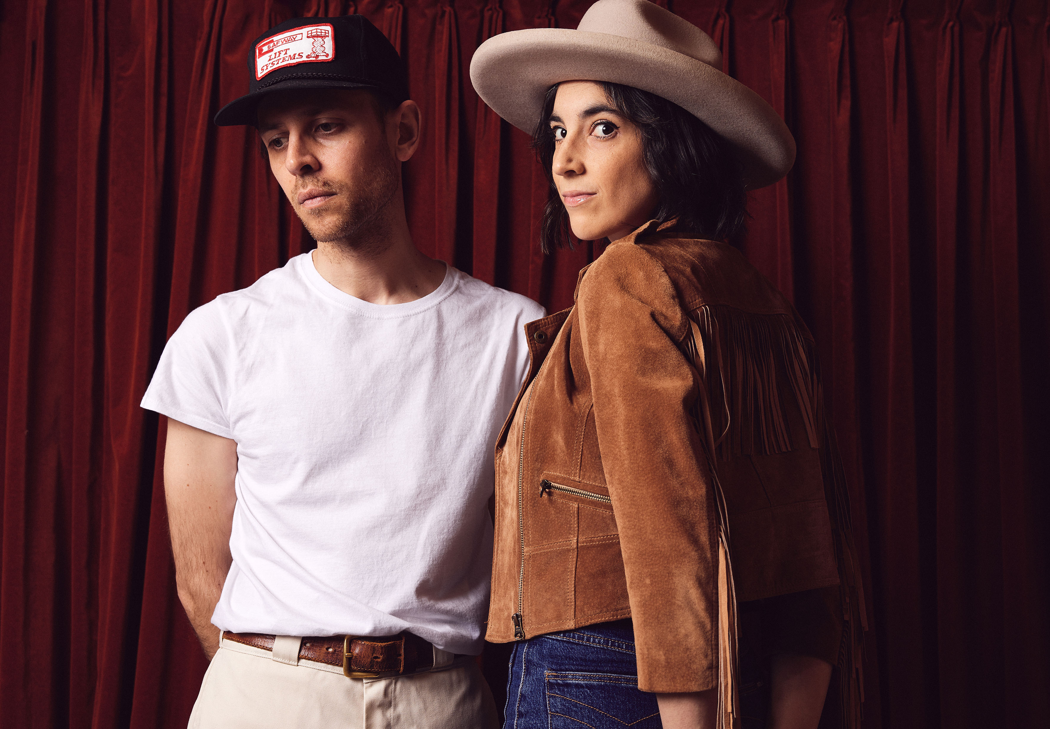 Um homem e uma mulher estão diante de uma cortina vermelha, o homem de boné e a mulher de chapéu de aba larga.