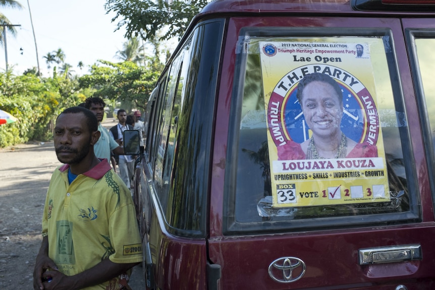 A man stands beside a car with an election cadidate poster taped to the back window.