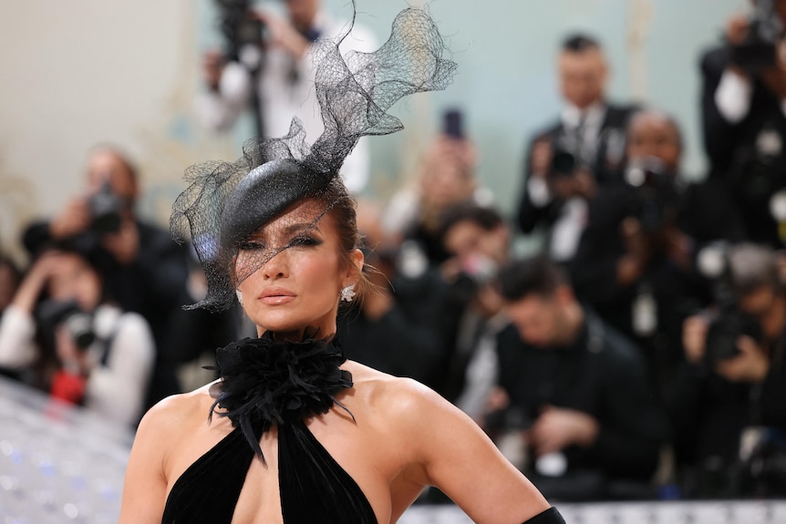 Jennifer Lopez poses at the Met Gala wearing a black fascinator and halter-neck dress in front of a crowd of paparazzi.