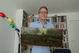A middle-aged white man with glasses stands in front of a bookshelf holding a Van Gogh painting