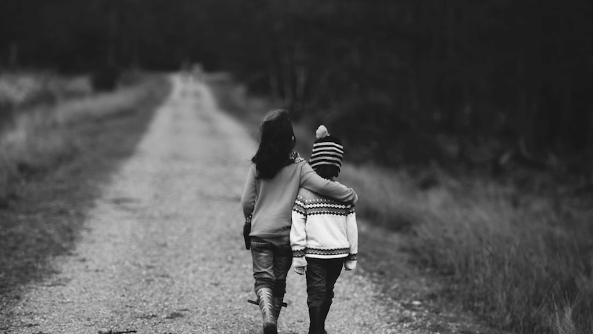 Black and White image of two children, a girl and boy, walking arm in arm down a long empty rural road. 