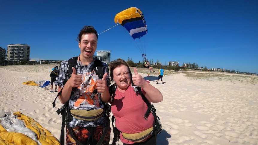 Photo of a young man and a woman  on a beach in skydiving gear after a jump grin and give the thumbs up to the camera.