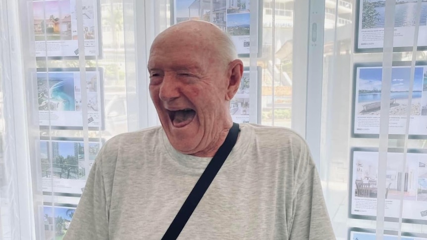 Denis, an elderly gentleman, stands in a grey t-shirt with a huge smile on his face.