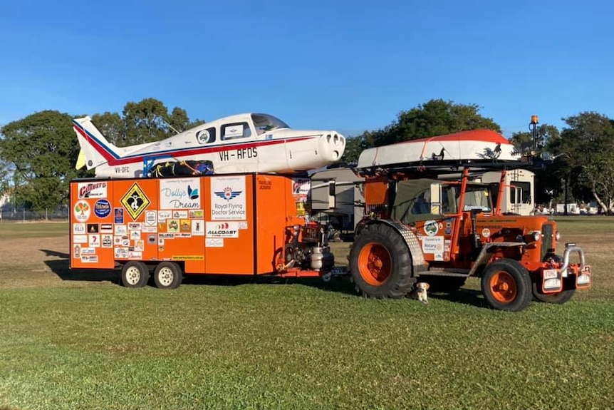 An orange tractor topped with a gyrocopter towing a trailer crowned with a Beechcraft Musketeer. 