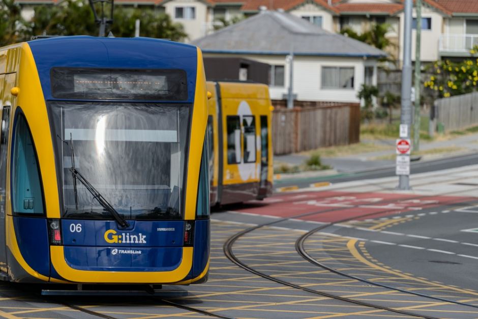 A带有 G:link 标志的蓝色和黄色电车转弯，白色和灰色，白色和赤陶屋顶的房子，背景是鲜花。” class=