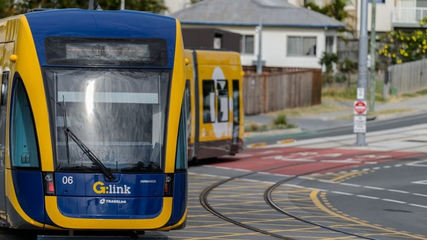 A blue and yellow tram with G:link logo turns, white and grey, white and terracotta roof houses, flowers in the background.