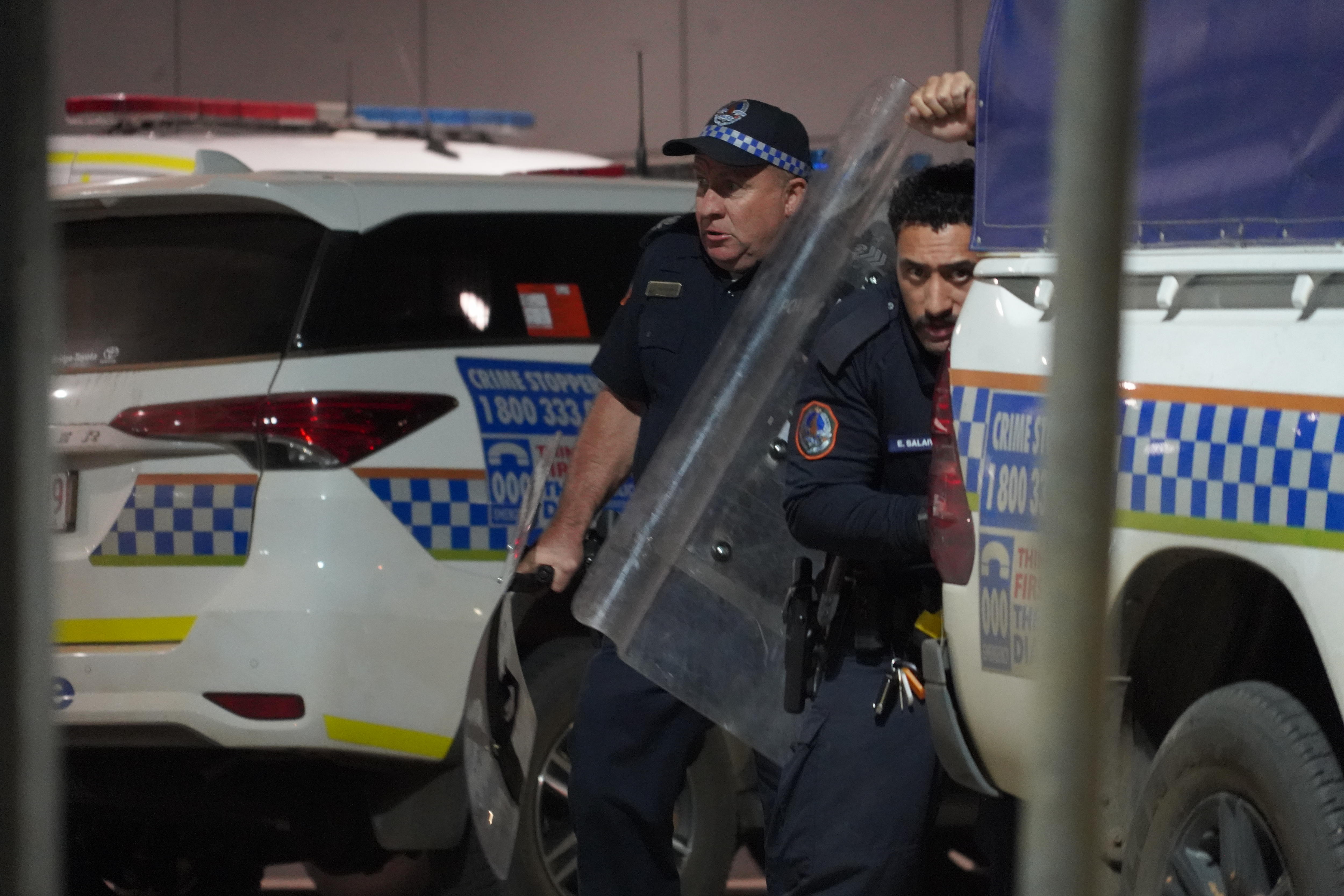 Dos policías con escudos protectores detrás de un vehículo policial.