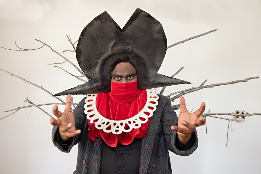 Man in red scarf covering face stares with arms out while wearing black headress