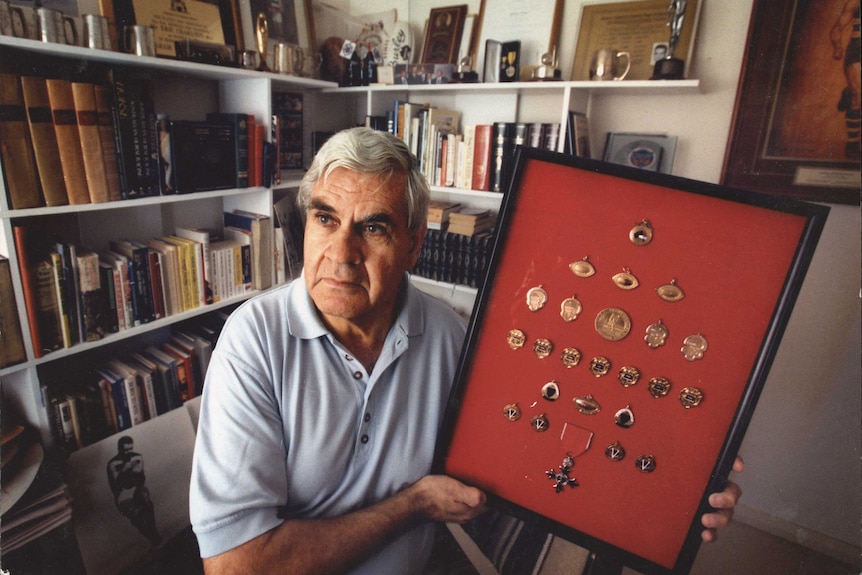 Graham 'Polly' Farmer holds up a frame with his best and fairest medals along with his MBE.