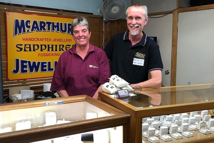 A man and woman stand at a jewellery shop front and smile at the camera.