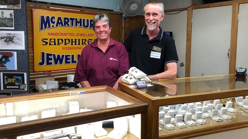 A man and woman stand at a jewellery shop front and smile at the camera.