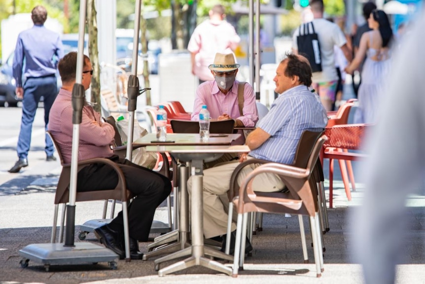 Tres hombres, uno con una máscara, se sientan en una mesa al aire libre en un café de Perth.
