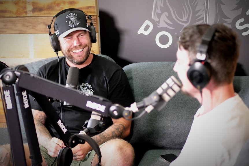 Two men on couches with headphones and microphones, one of them is clearer in the image and is smiling