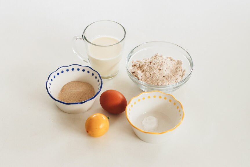 Ingredients for pikelets, including flour, sugar, milk and eggs, assembled in separate small bowls on a bench.