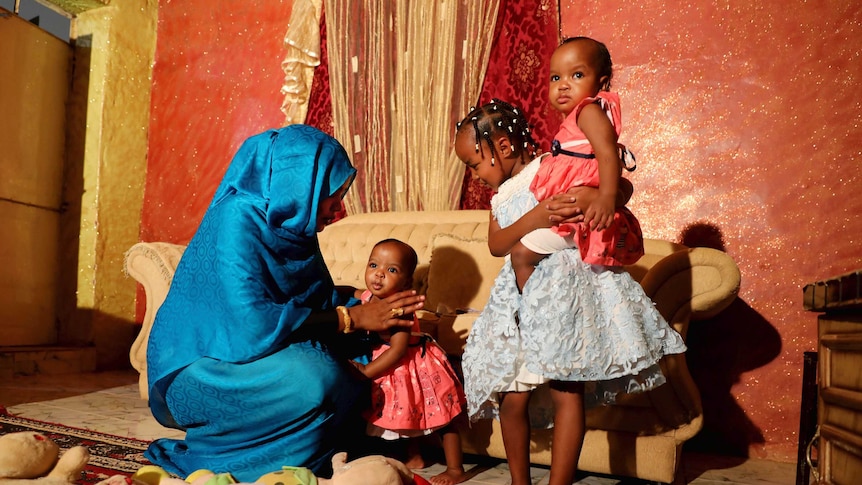A mother plays with her daughters in their home as toys are seen on the floor.