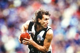 A colour photo of an AFL footballer running with the ball, crowd out-of-focus in background