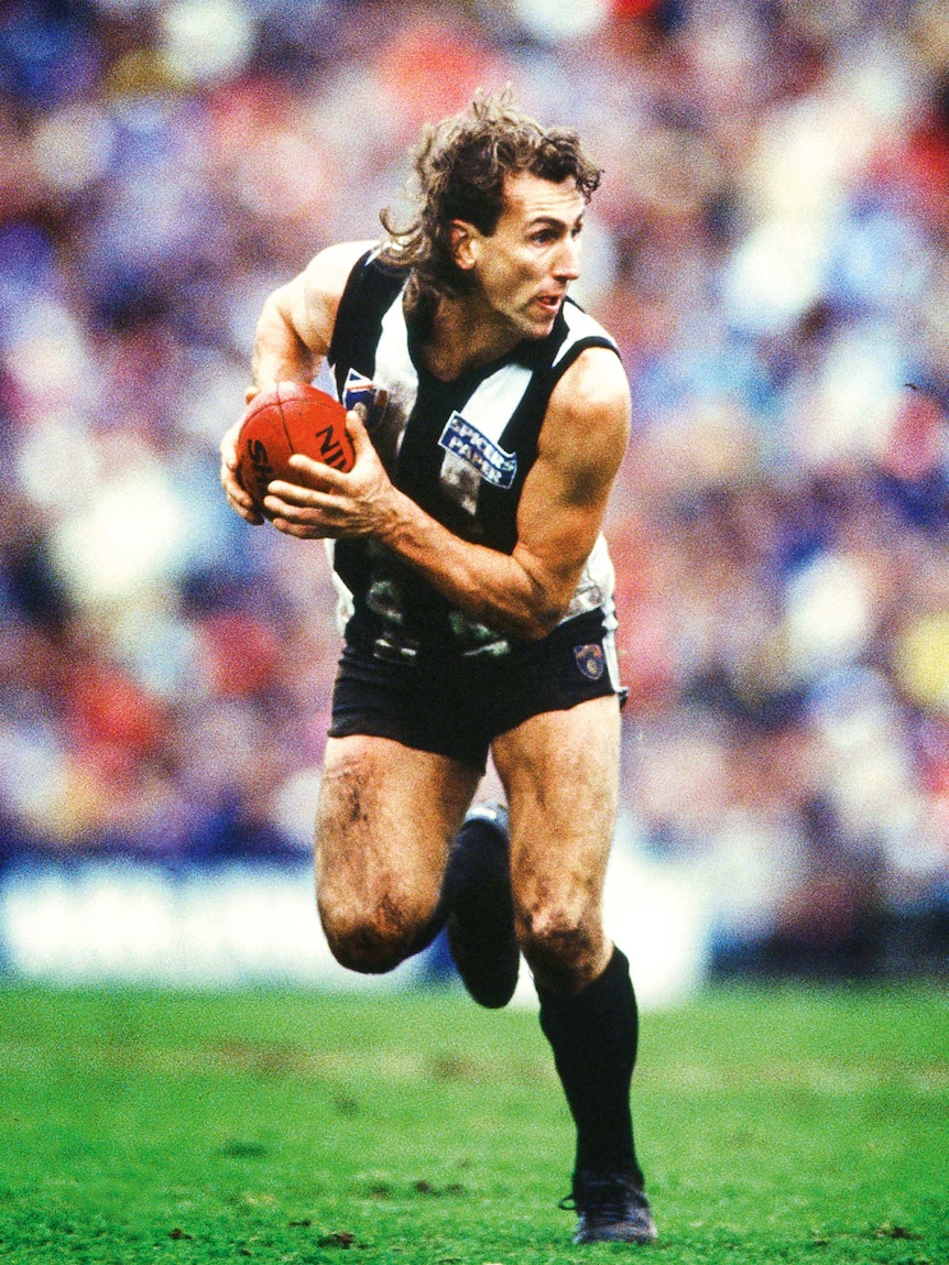 A colour photo of an AFL footballer running with the ball, crowd out-of-focus in background