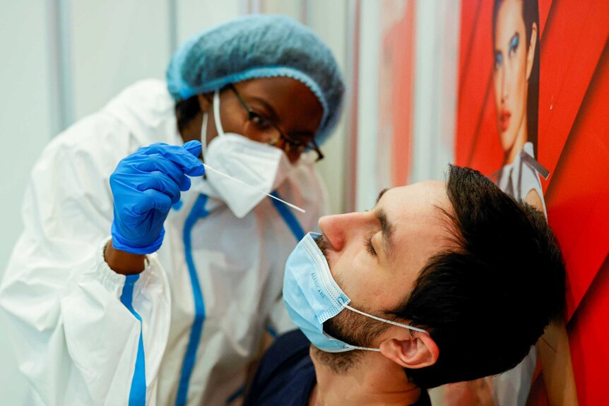 A health worker in PPE holds a long swab just above a man's nose