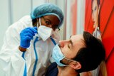 A health worker in PPE holds a long swab just above a man's nose