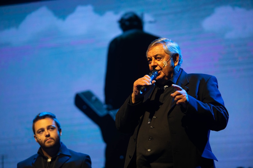 A man sings against a projection of Johnny Cash with guitar. Another singer looks on.