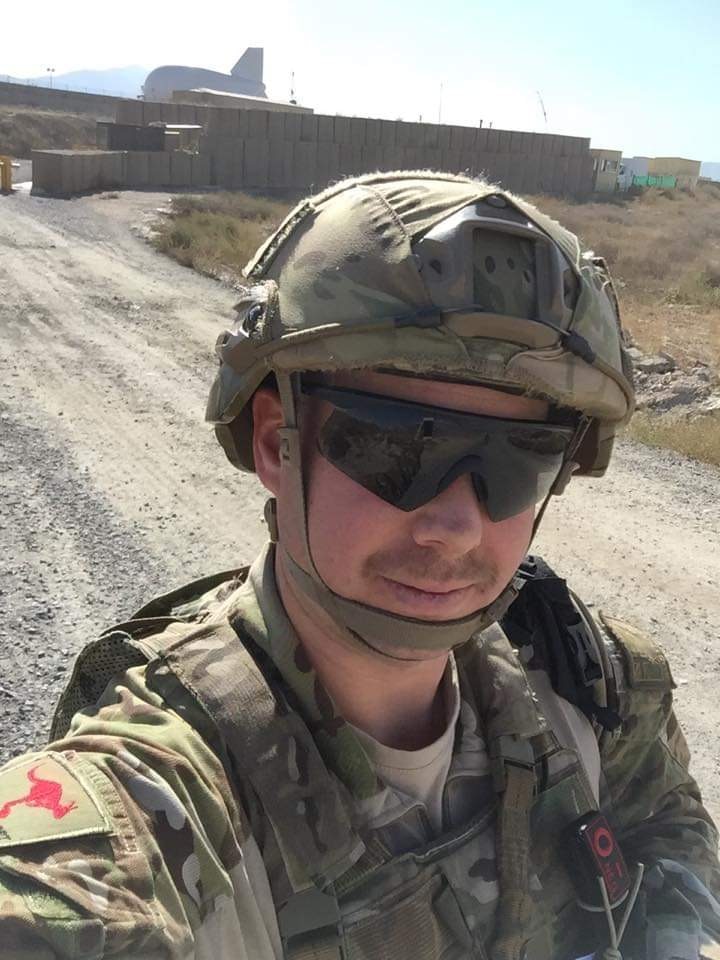 A man wearing a camouflage military uniform and helmet standing on a sandy road smiles for a selfie.