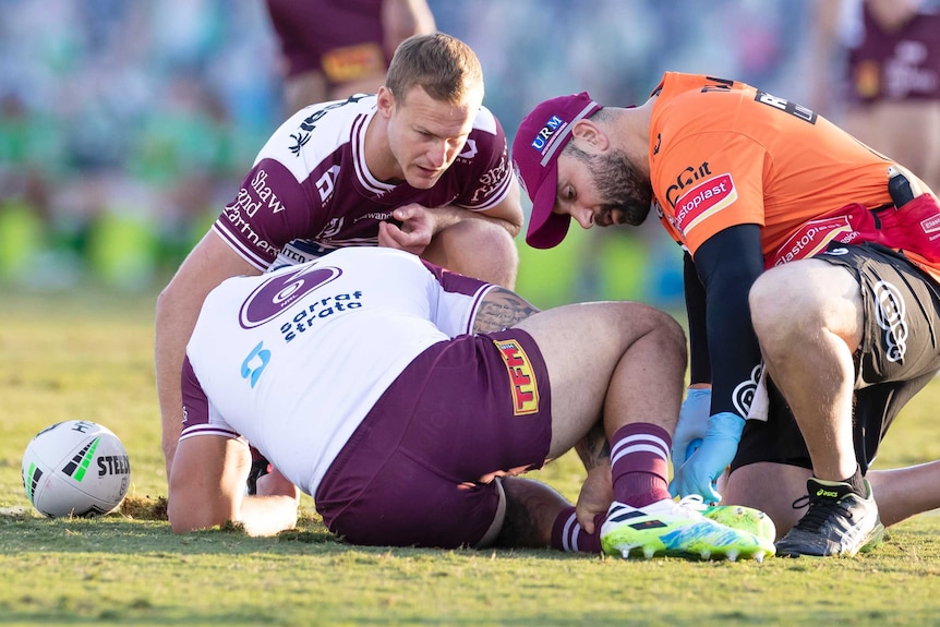 An NRL player lies on the ground injured as his teammate and a trainer check on him during a game.