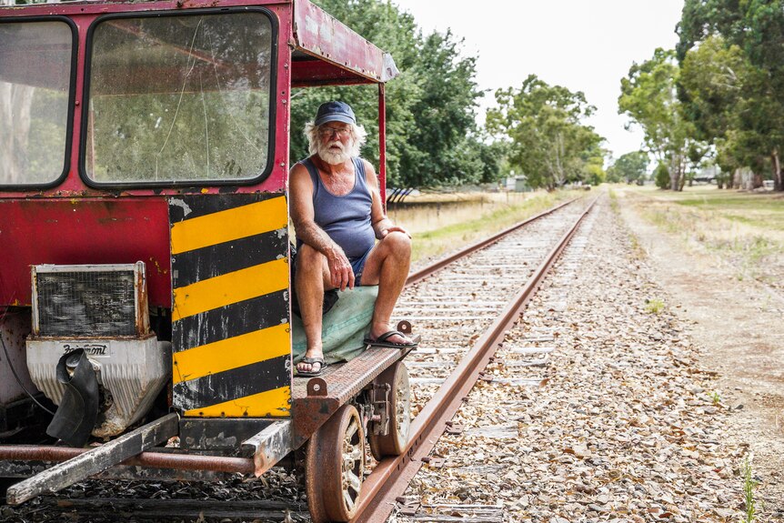 A man sits on a rusty red open railway carriage, trees behind him.