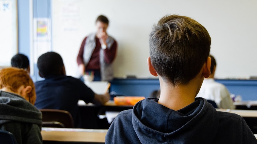 A boy faces a teacher who is standing in front of a blackboard.