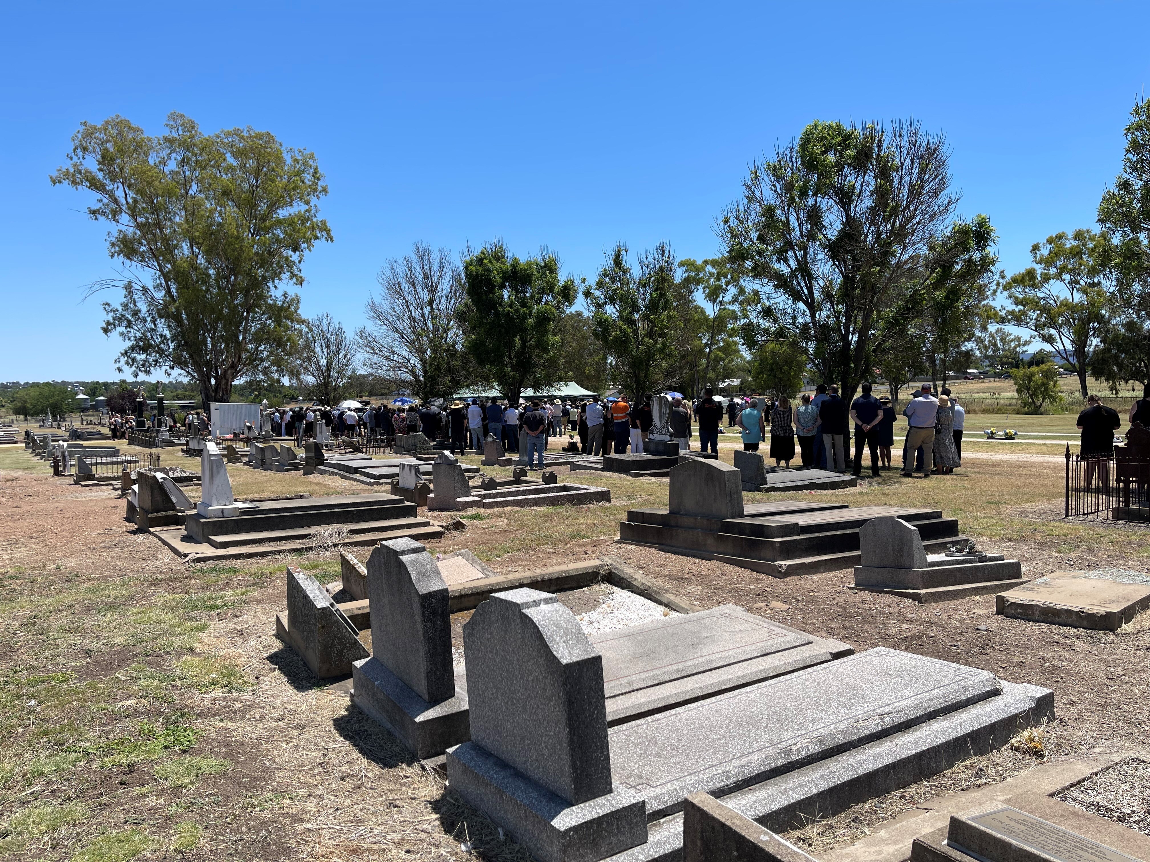 A crowd of people at a cemetery with headstones in the foreground and trees