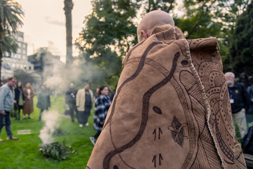 Man wrapped in traditional dress featuring Indigenous markings, with smoking fire in background