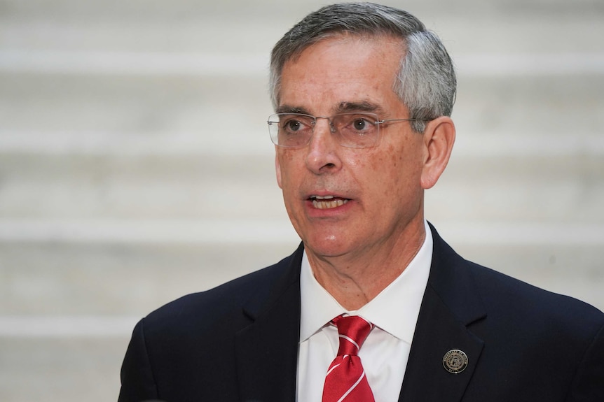 Brad Raffensperger, wearing a suit, speaks in front of a white background.