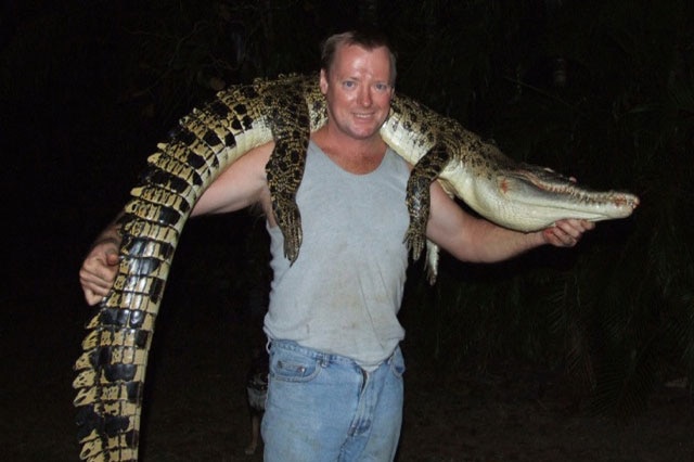 A man in a grey singlet holds a dead crocodile draped over his shoulders.
