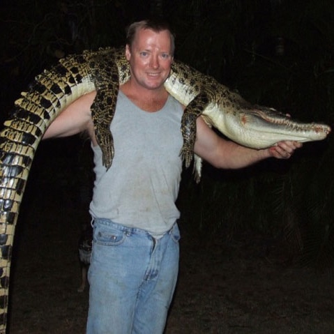 A man in a grey singlet holds a dead crocodile draped over his shoulders.