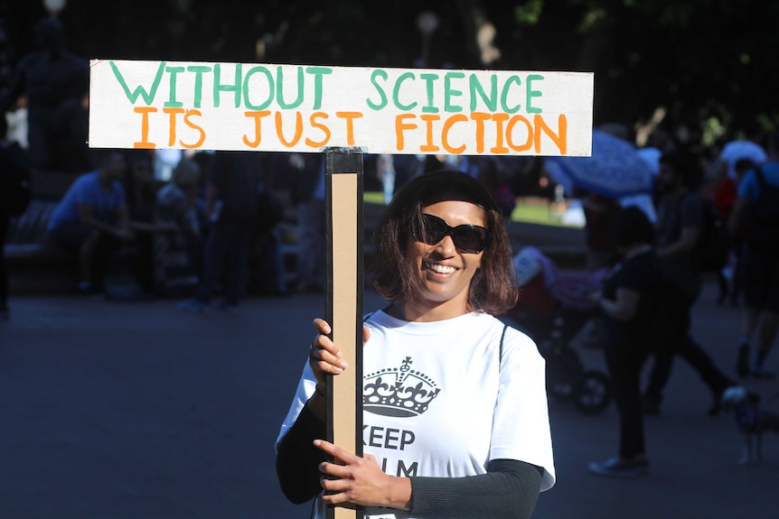 A woman wearing a "keep calm" t-shit holds a sign reading: "Without science it's just fiction"