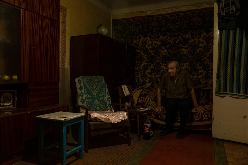 An old man sits on a bed in a small apartment. There is a chair, small table and wardrobe next to him