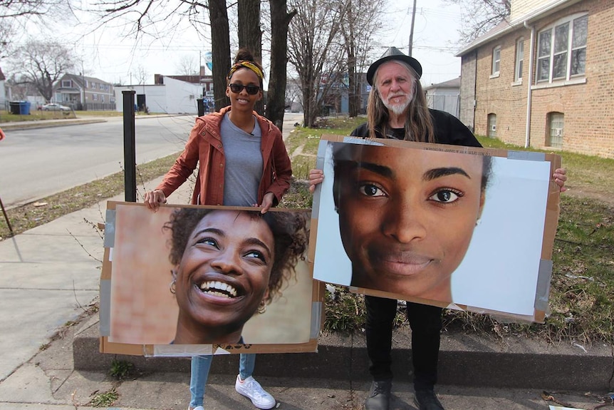 George Gittoes and a female friend hold large photos of women's faces in suburban Chicago.