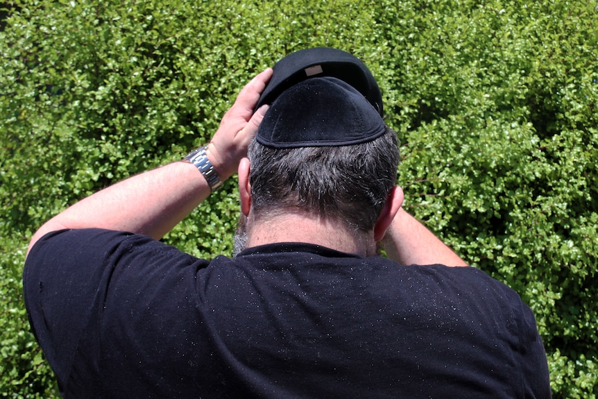 A man puts a cap over his Jewish Kippah while facing a bush, with the camera behind him.