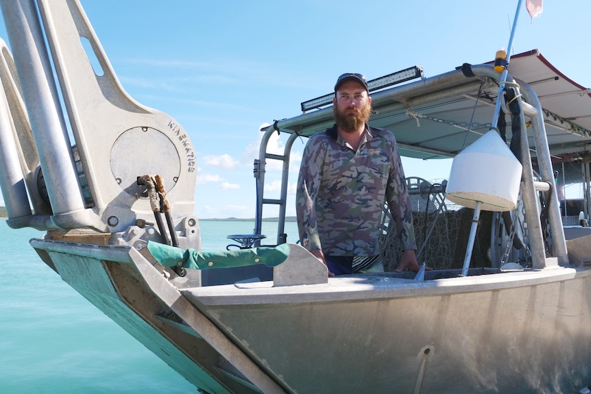 A man stands on a commercial fishing boat with blue sky above him and blue water beneath him 