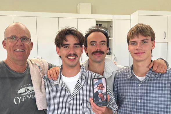 Three young men stand with their father, one holding a phone which they're using to FaceTIme their sister