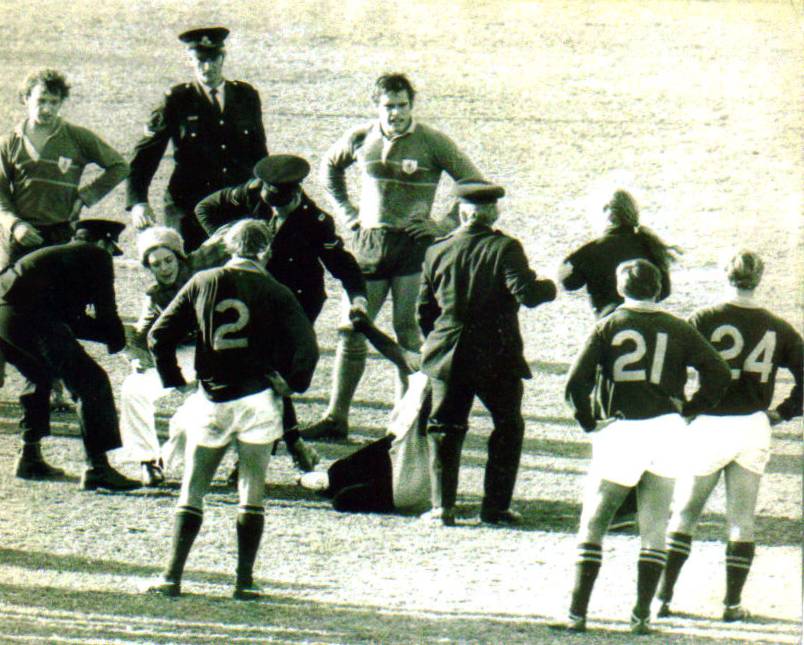 A group of men, and a police officer arresting a woman as players in shorts and numberd jersey look on with hands on hips.