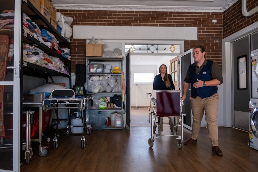 A man and a woman push a coffin on a stand through a garage