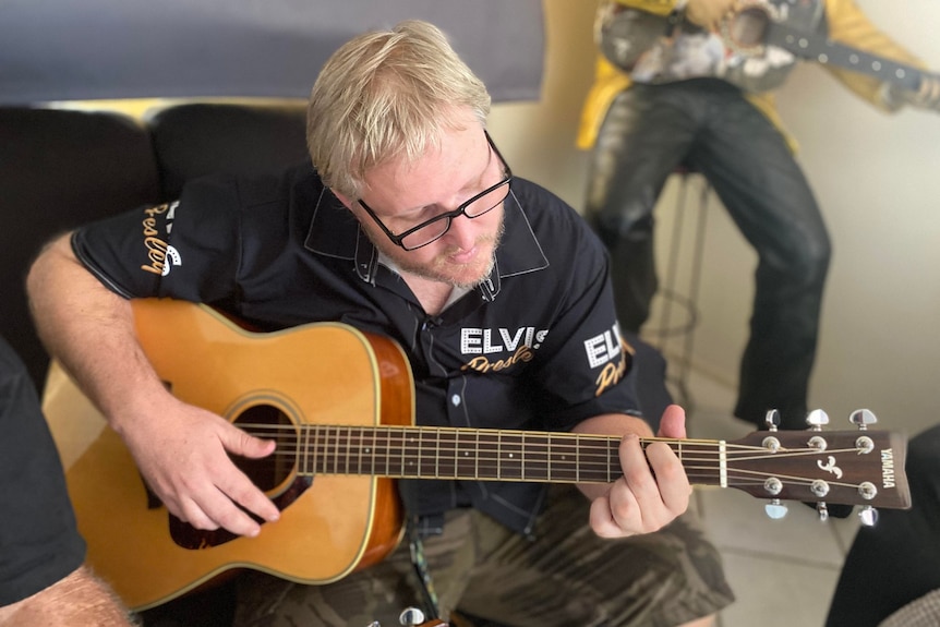 Joshua Clarke seated on a couch playing a guitar, an Elvis Presley statue is visible behind him