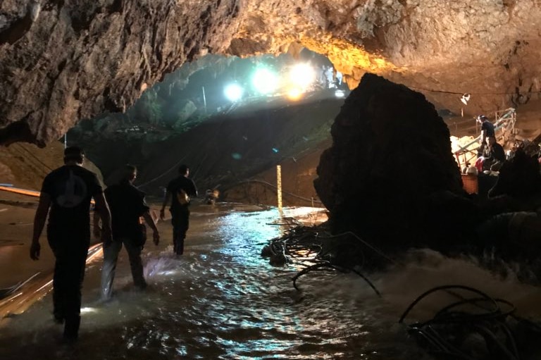 View inside a section of cave with a high ceiling, as four people walk through shallow waters. There are lights in the cave.