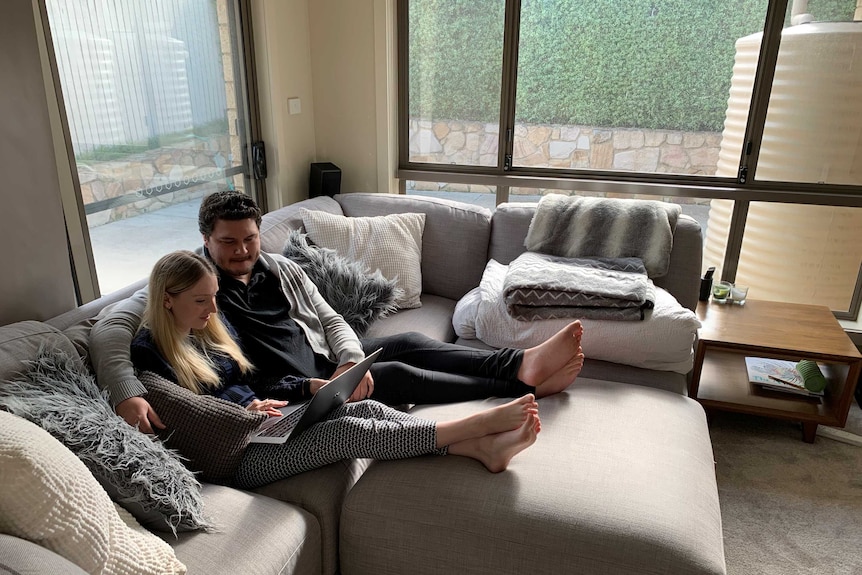 The couple sits on their couch looking at a laptop.