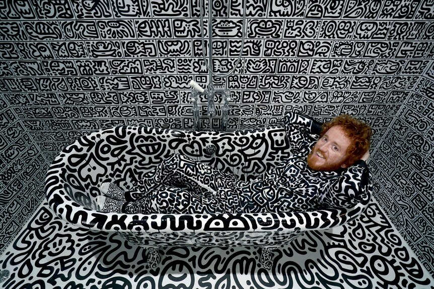 Sam Cox lays in a bathtub. The entire room, including his clothes, is covered in black and white art. 