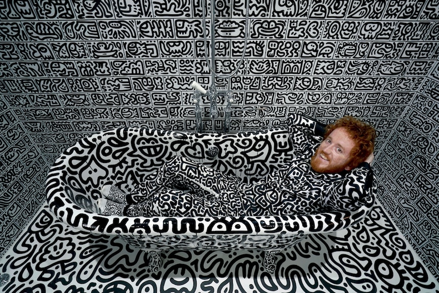 Sam Cox lays in a bathtub. The entire room, including his clothes, is covered in black and white art. 