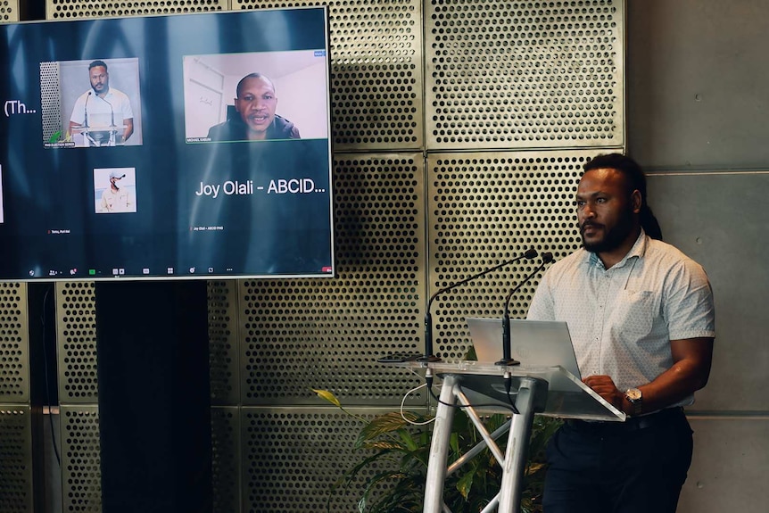 A PNG man stands behind a lectern and to his side a screen showing two more men in video conference.